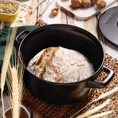Loaf of bread in a black pot on a rustic wooden table with grains and seeds.