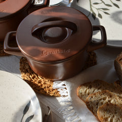 Brown ceramic pot with lid on a textured surface, surrounded by bread and plates.