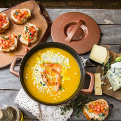 Yellow soup with shrimp in a black bowl on a wooden table with bread and cheese.