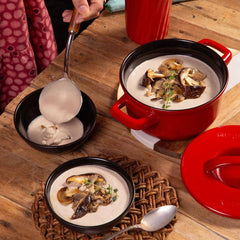 Red pot with mushroom soup on a wooden table, surrounded by spoons and a person's hand.